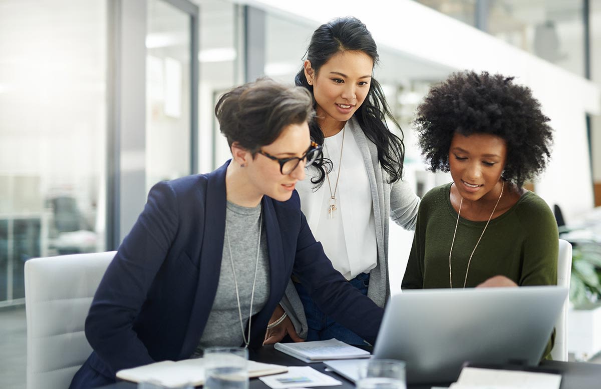 Three women in a professional office reviewing work on a laptop computer